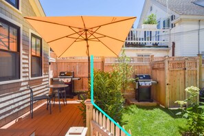 Un jardin avec une terrasse en bois, un barbecue, une table de patio avec des chaises et un grand parasol.