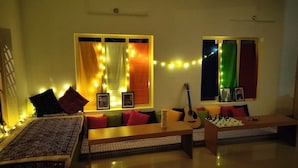 A cozy living room with a sofa, a coffee table, and a guitar. There are two windows with curtains and string lights. - The Hostel Stories Varkala (Varkala)