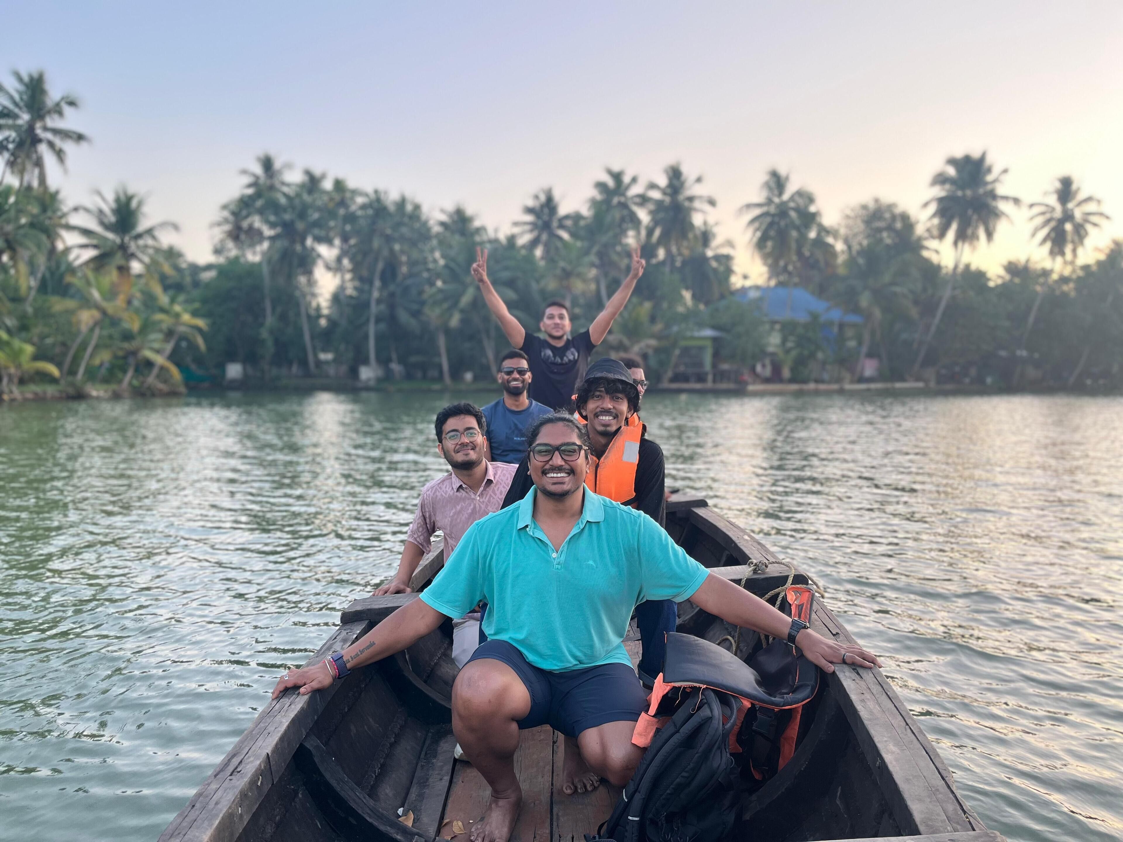 A group of people on a wooden boat with a calm water body and palm trees in the background.