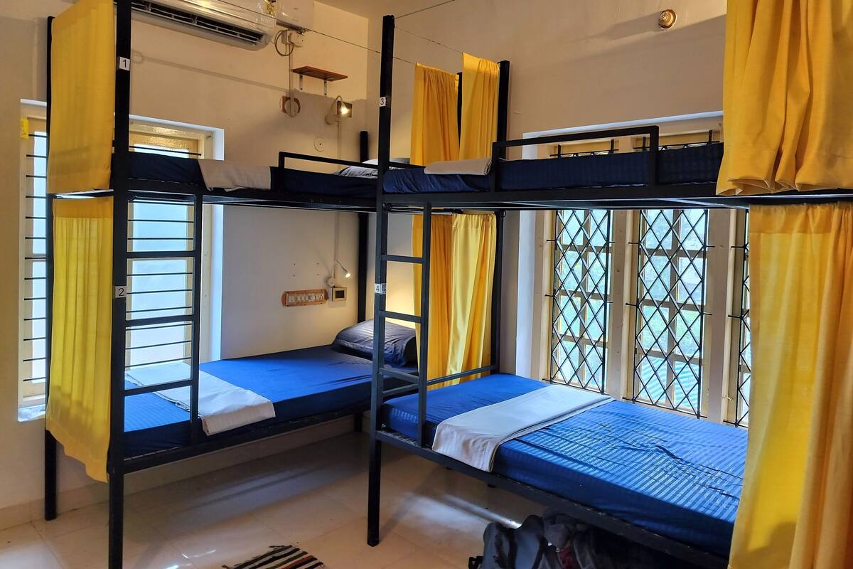 A dormitory room with bunk beds, a window with yellow curtains, and an air conditioning unit.