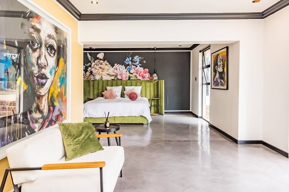 A modern bedroom with a green upholstered headboard, a white bed with pink and red pillows, a large floral artwork on the wall, and a white sofa with a green cushion.