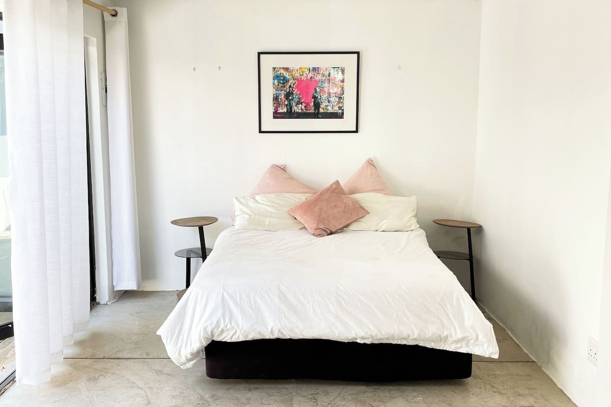 A bedroom with a bed, a framed artwork, two bedside tables, and a view of the outside.