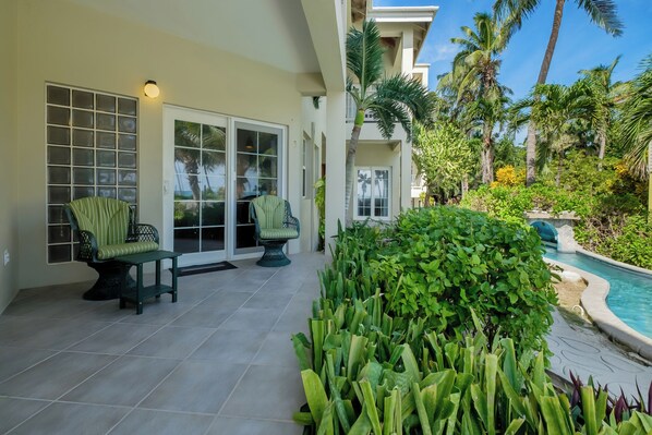 A covered patio with two chairs, a pool, and lush greenery.
