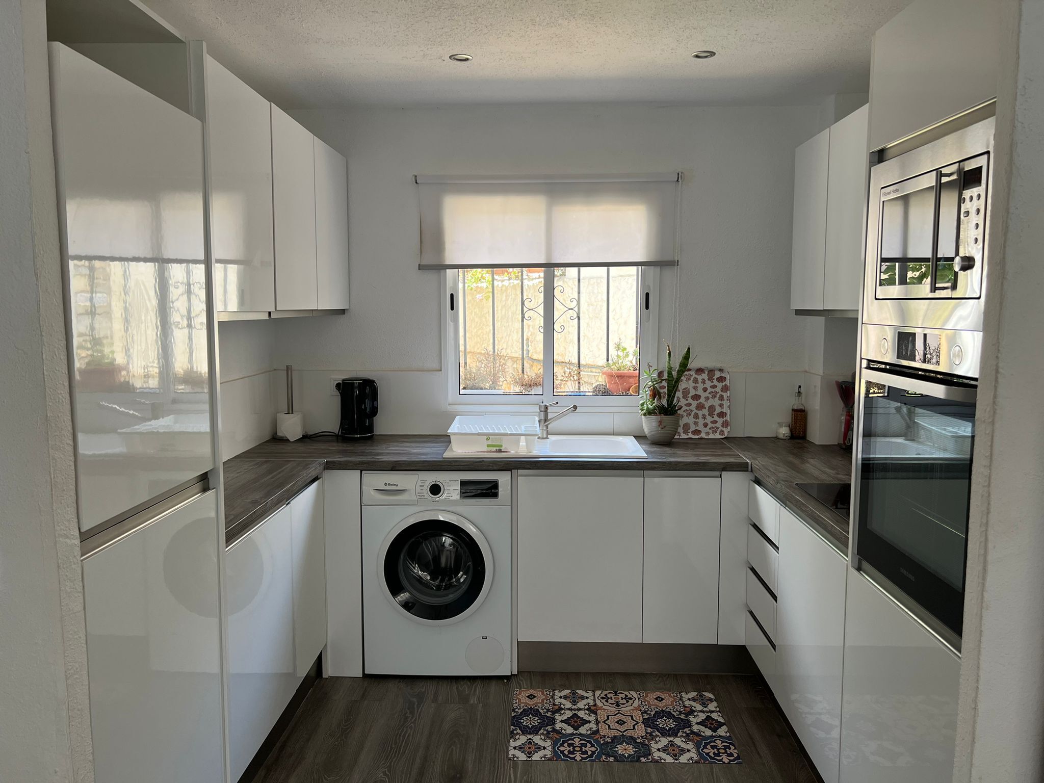 A compact kitchen with a built-in washer and dryer, a window with blinds, and a stainless steel oven.