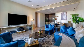 Een moderne woonkamer met een flatscreen-tv, blauwe banken, een salontafel met fruit en een eethoek op de achtergrond.