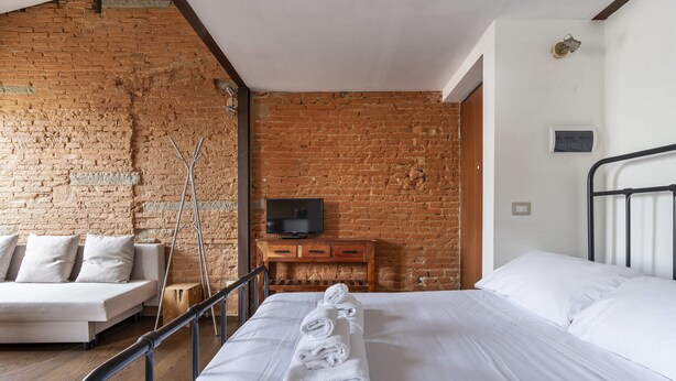 A hotel room with a brick wall, a bed with white linens, a wooden nightstand with a TV, and a sofa.
