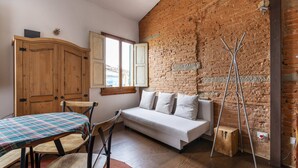 A room with a wooden wardrobe, a sofa, a dining table with a plaid tablecloth, and a brick wall.