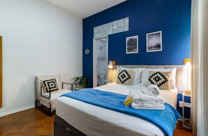 Une chambre d’hôtel comprenant un lit, une chaise, une table de chevet, un miroir et un mur orné de deux tableaux encadrés.