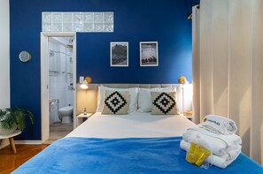 Une chambre à coucher avec un lit, deux oreillers, une table de chevet, une horloge murale et une salle de bain visible à travers une porte ouverte.