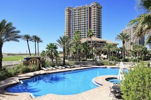 A swimming pool surrounded by palm trees and lounge chairs, with a tall hotel building in the background.