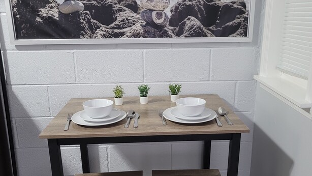 Un coin repas avec une table en bois dressée pour deux personnes, un banc et un cadre offrant une vue sur l’océan.