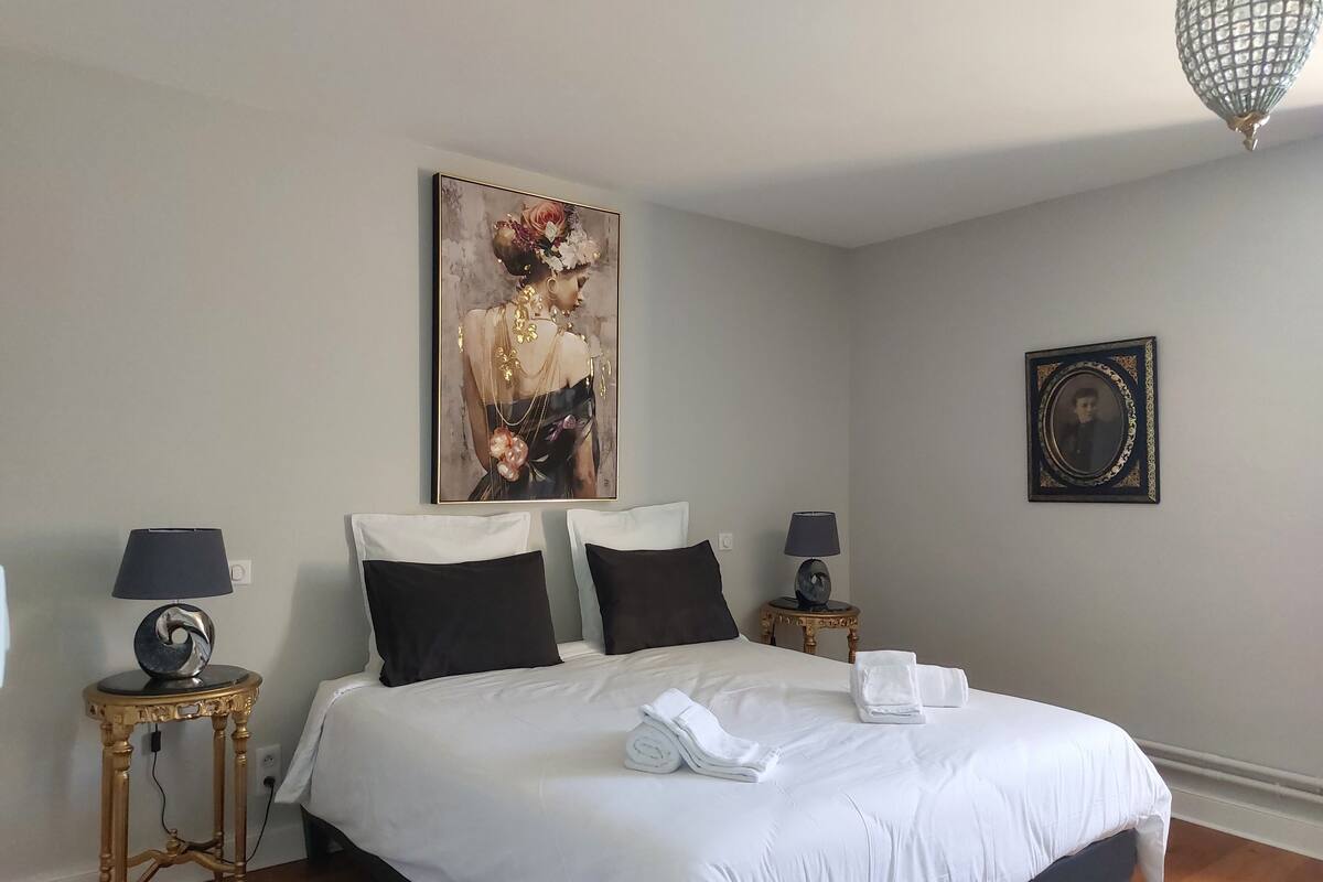 Una cama bien hecha con sábanas blancas, dos mesitas de noche con lámparas, un retrato enmarcado en la pared y un cuadro sobre la cama.