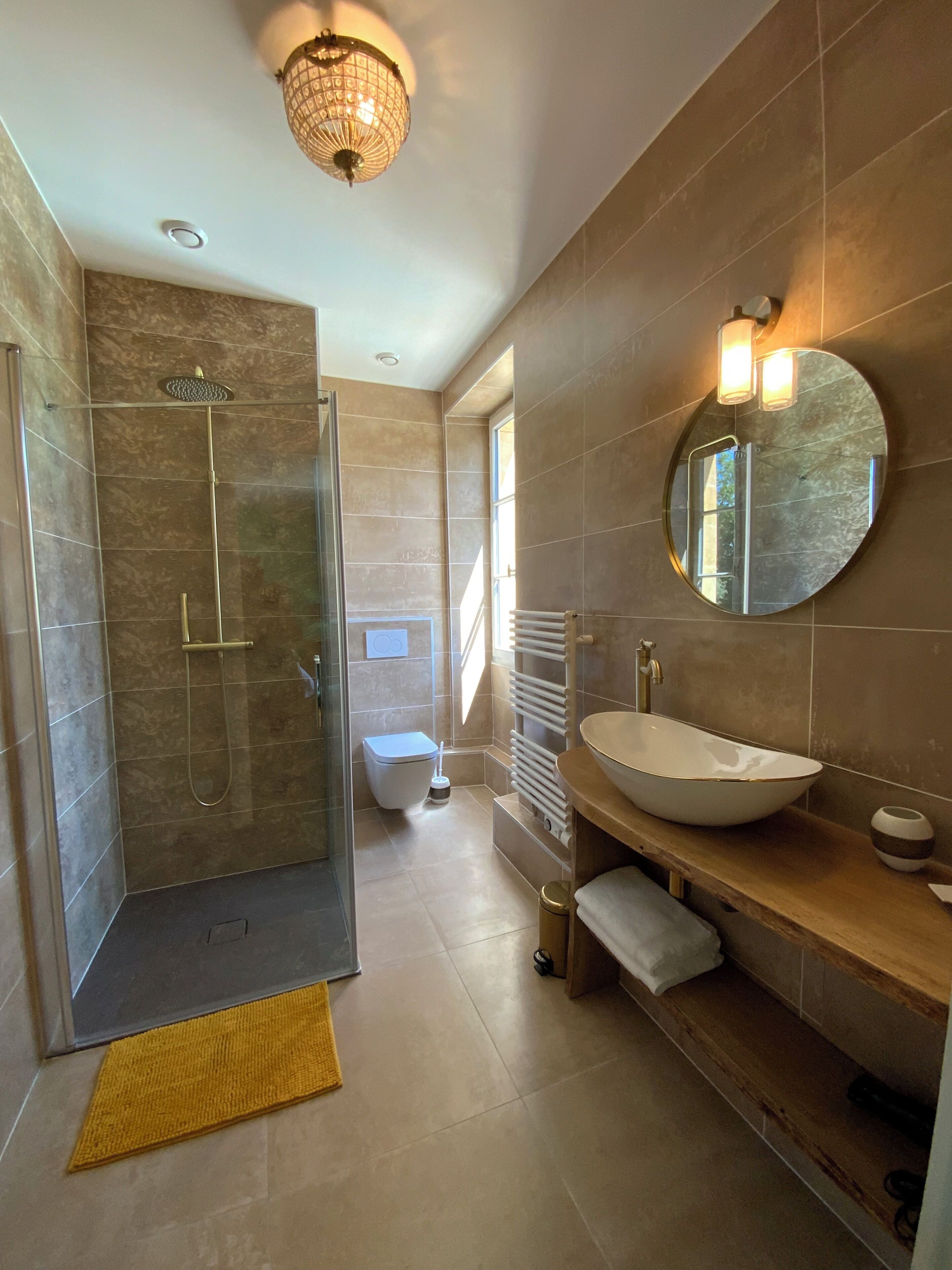 Un baño moderno con ducha envuelta en vidrio, espejo redondo, lavamanos blanco y suelo de baldosas color beige.