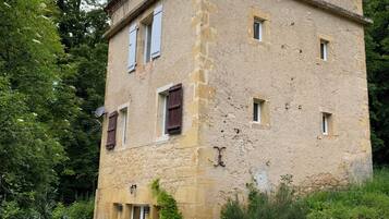 Un bâtiment rustique en pierre, doté de nombreuses fenêtres et d’une porte, entouré d’une végétation luxuriante et accessible par un escalier en pierre.