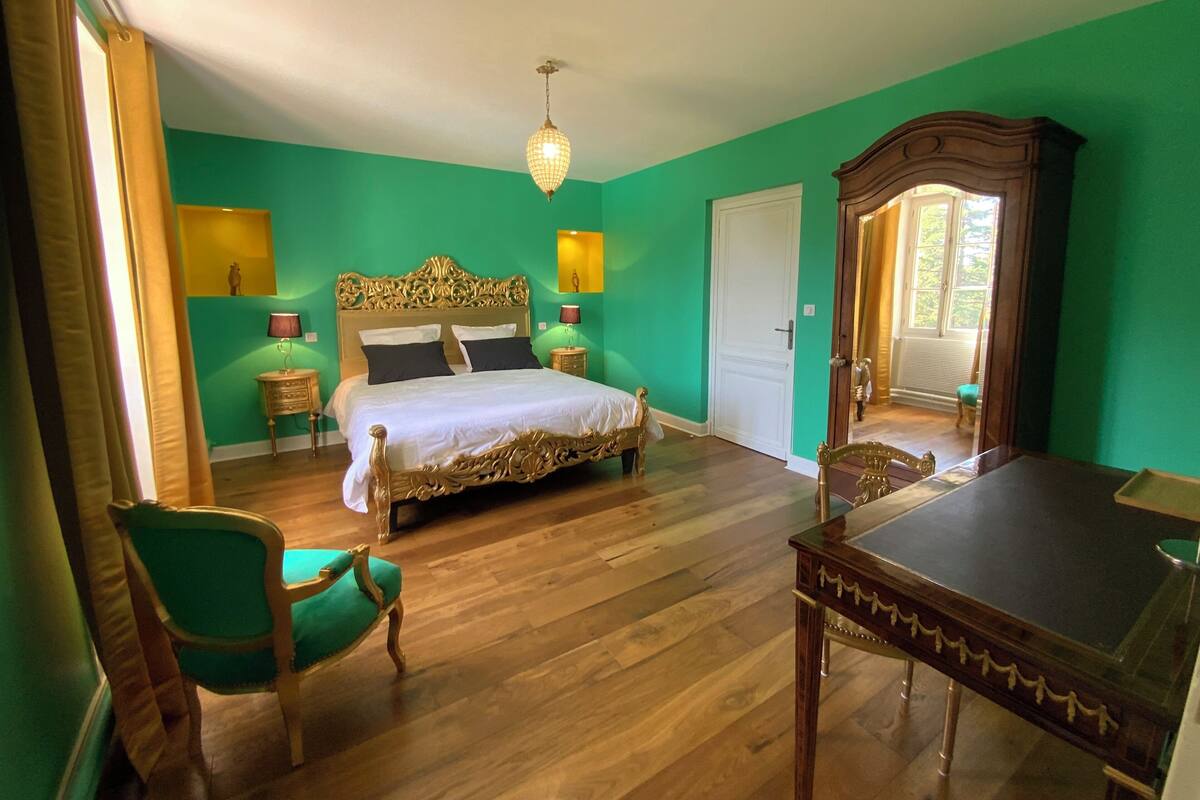 Una habitación con una cama grande, una mesa de madera, una silla verde y un espejo.