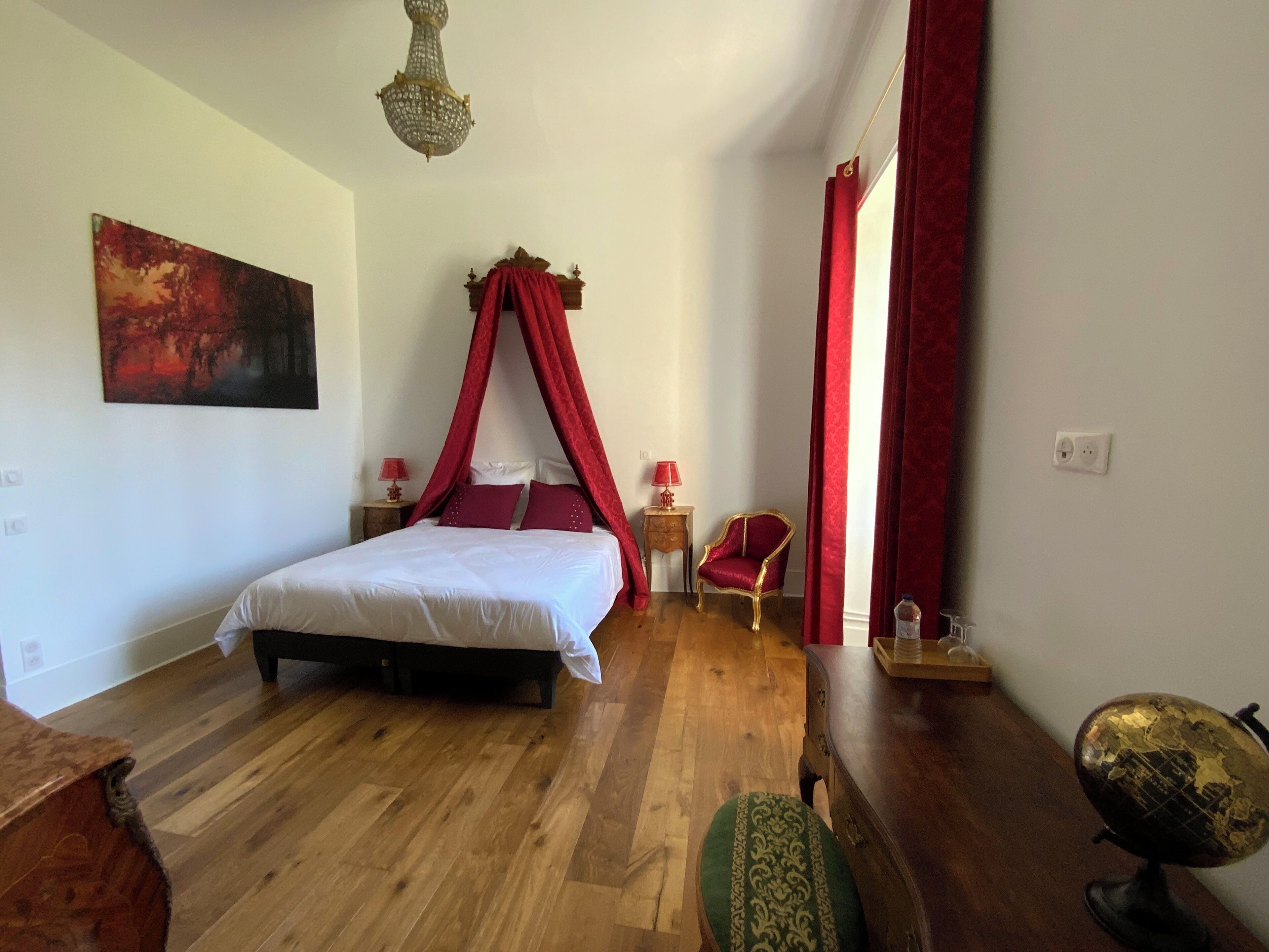 Un dormitorio con cama con dosel, suelo de madera, dosel rojo, un globo terráqueo y una silla.