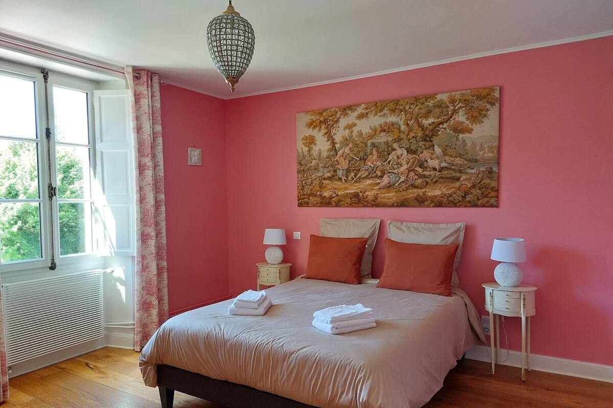 Un dormitorio con cama, dos mesitas de noche con lámparas, un radiador, una ventana con cortinas y un cuadro de paisaje en la pared.