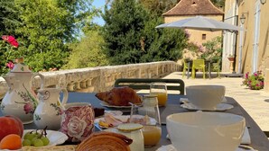 Desayuno al aire libre con croissants, frutas y bebidas sobre una mesa, con vista a un jardín y un edificio con tejado de azulejos.