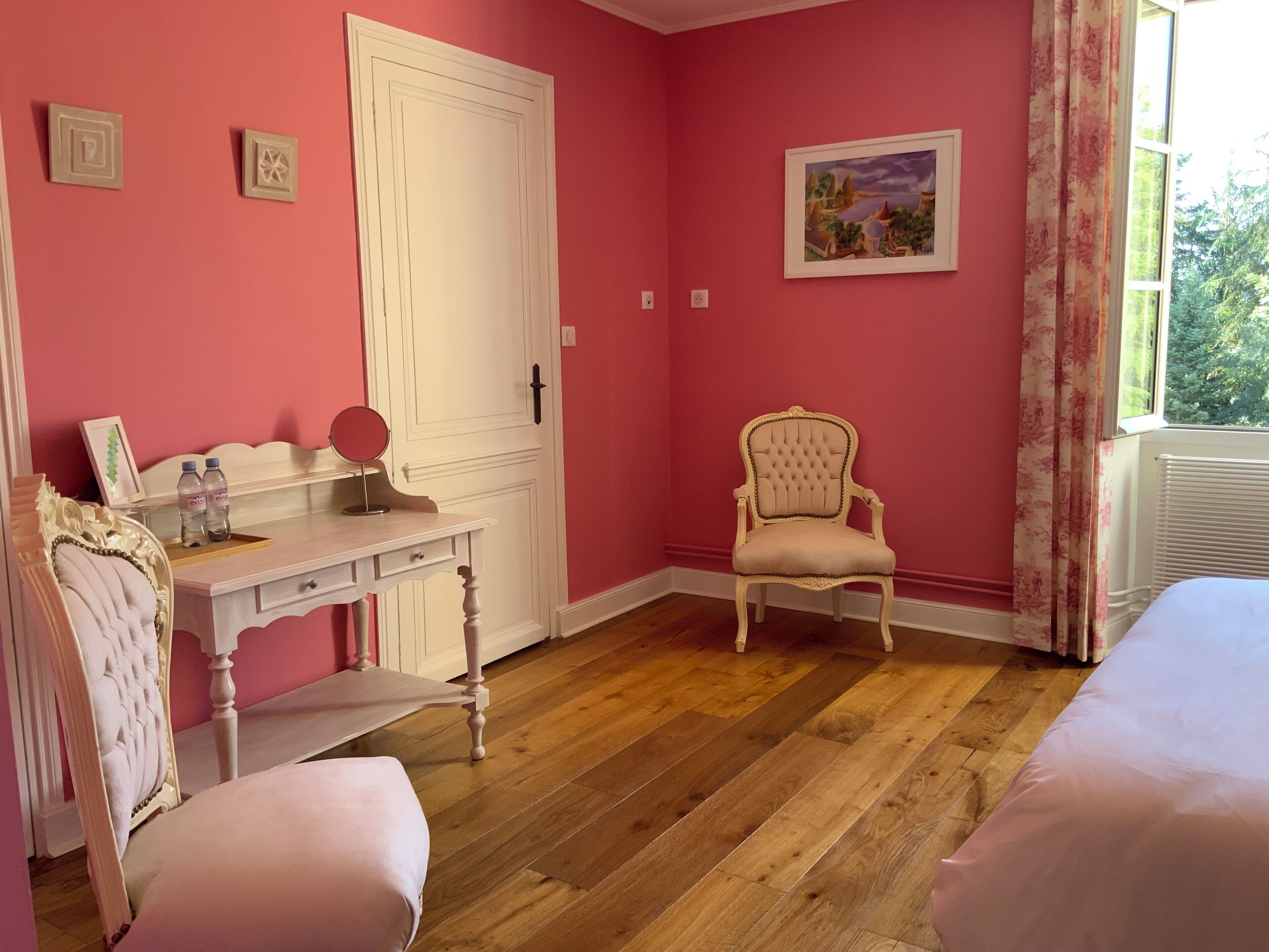 A room with pink walls, a white door, a vanity with a mirror, two pink chairs, and a bed.
