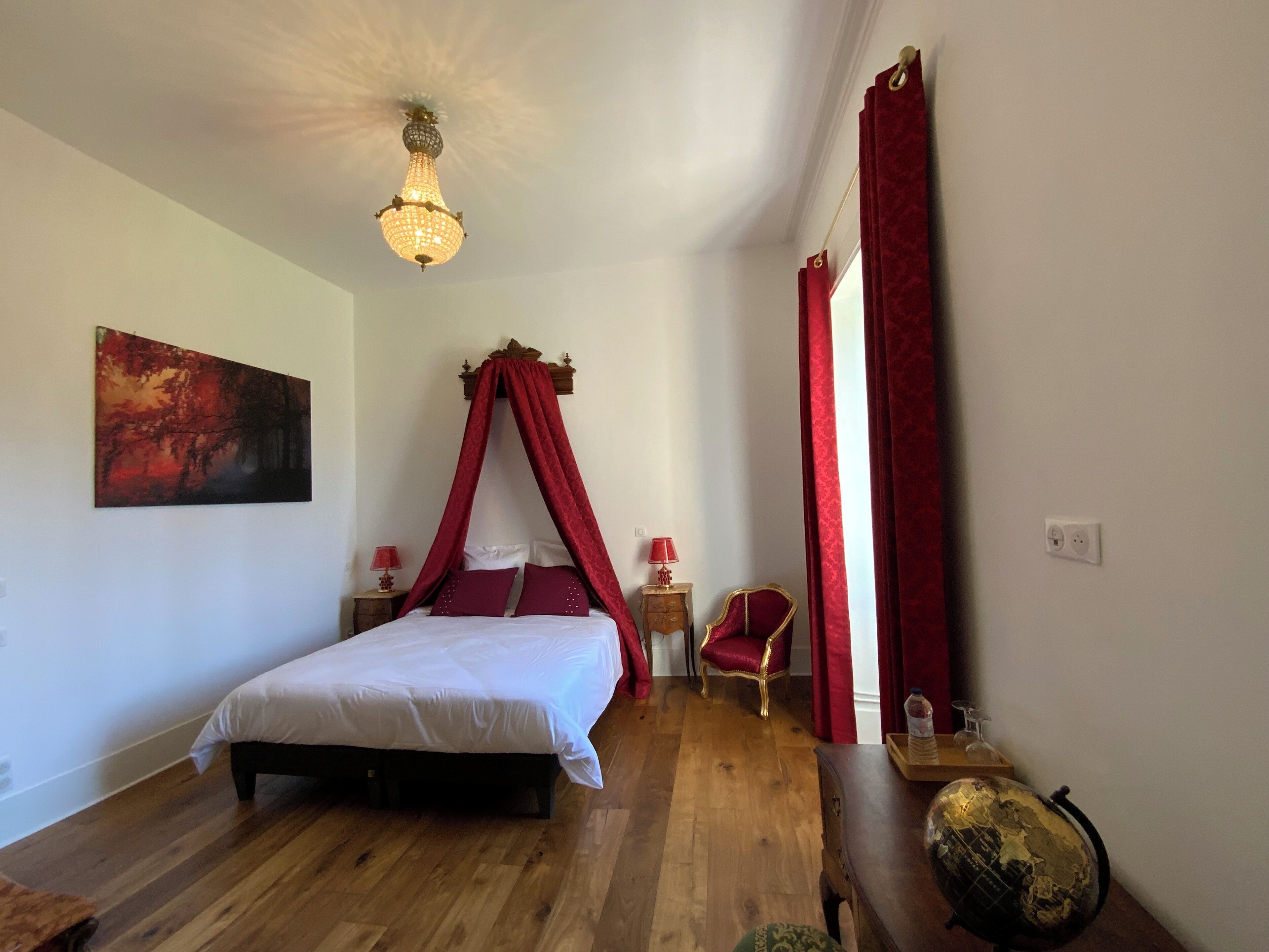 A bedroom with a four-poster bed, a chandelier, a red canopy, a globe, and a painting on the wall.