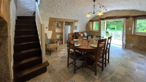 Un comedor rústico con una mesa de madera dispuesta para cuatro personas, rodeada de sillas. Una escalera conduce a un nivel superior. La habitación presenta paredes de piedra a la vista y un techo abovedado con luces colgantes.