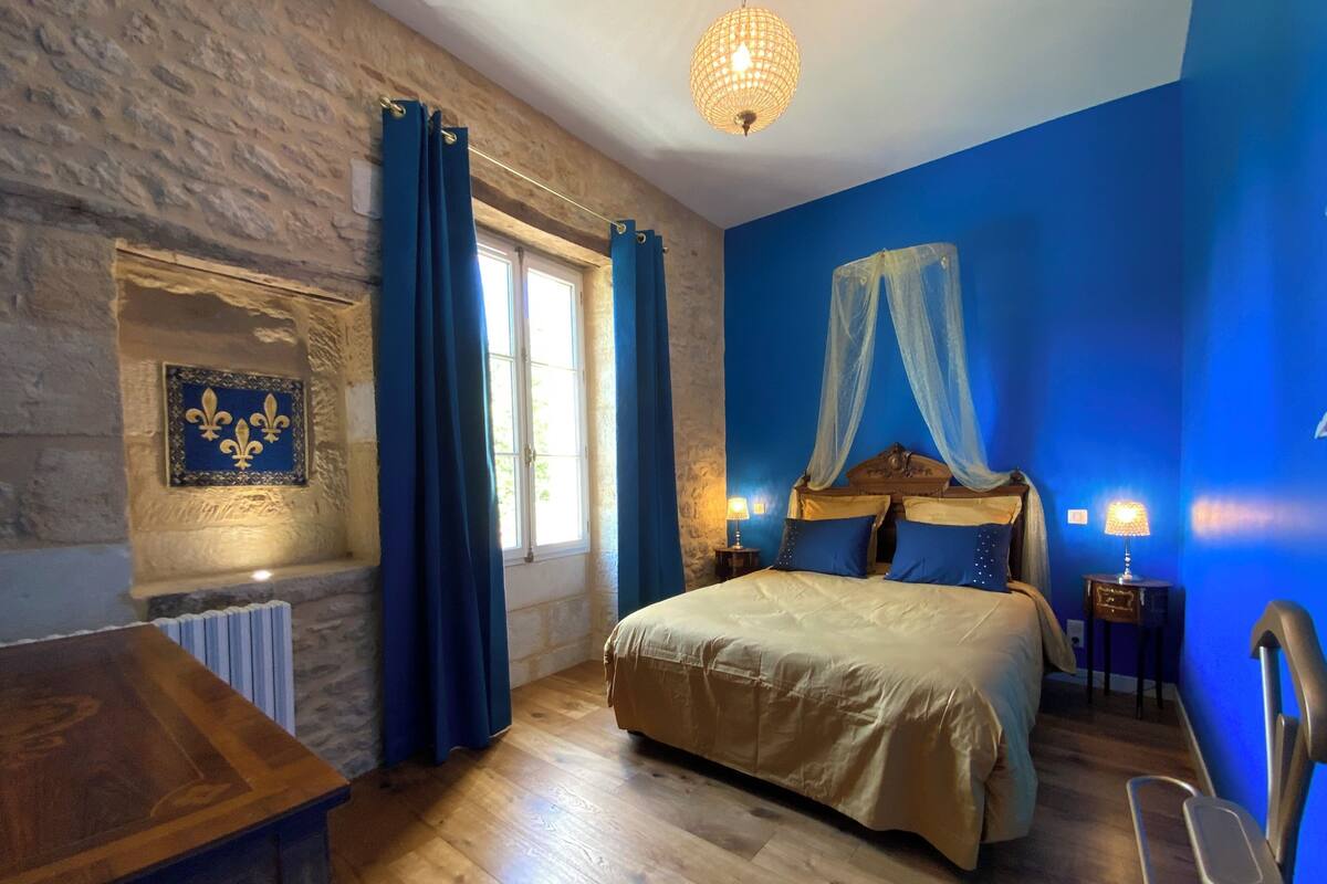 Un dormitorio con pared de piedra, cama de madera con ropa de cama azul, mesita de noche con lámpara y ventana con cortinas azules.