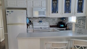 Une cuisine avec des meubles blancs, un réfrigérateur, une cuisinière et deux tabourets de bar en osier.