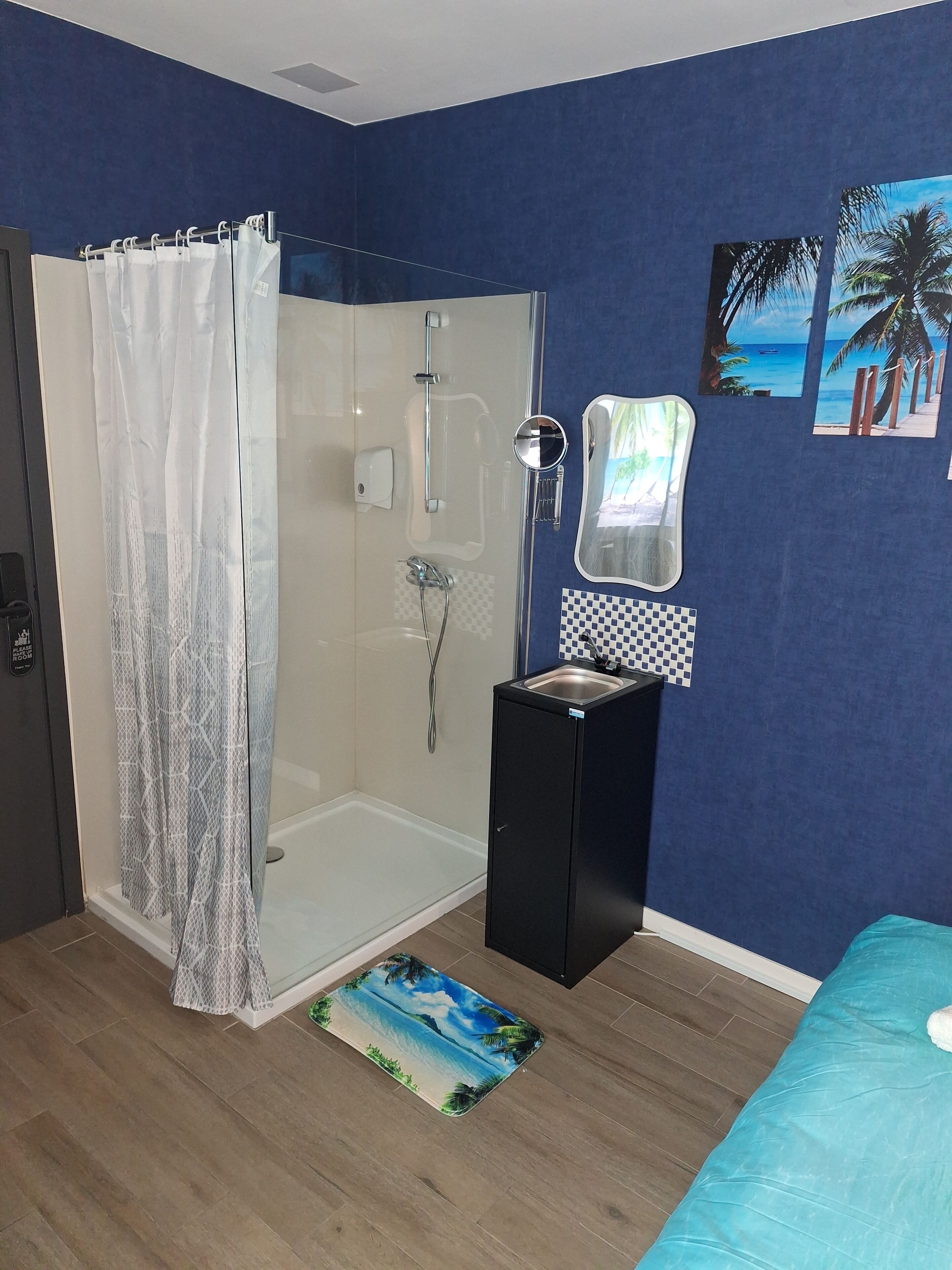 A bathroom with a glass shower enclosure, a sink with a mirror, and a bed with a blue bedspread.