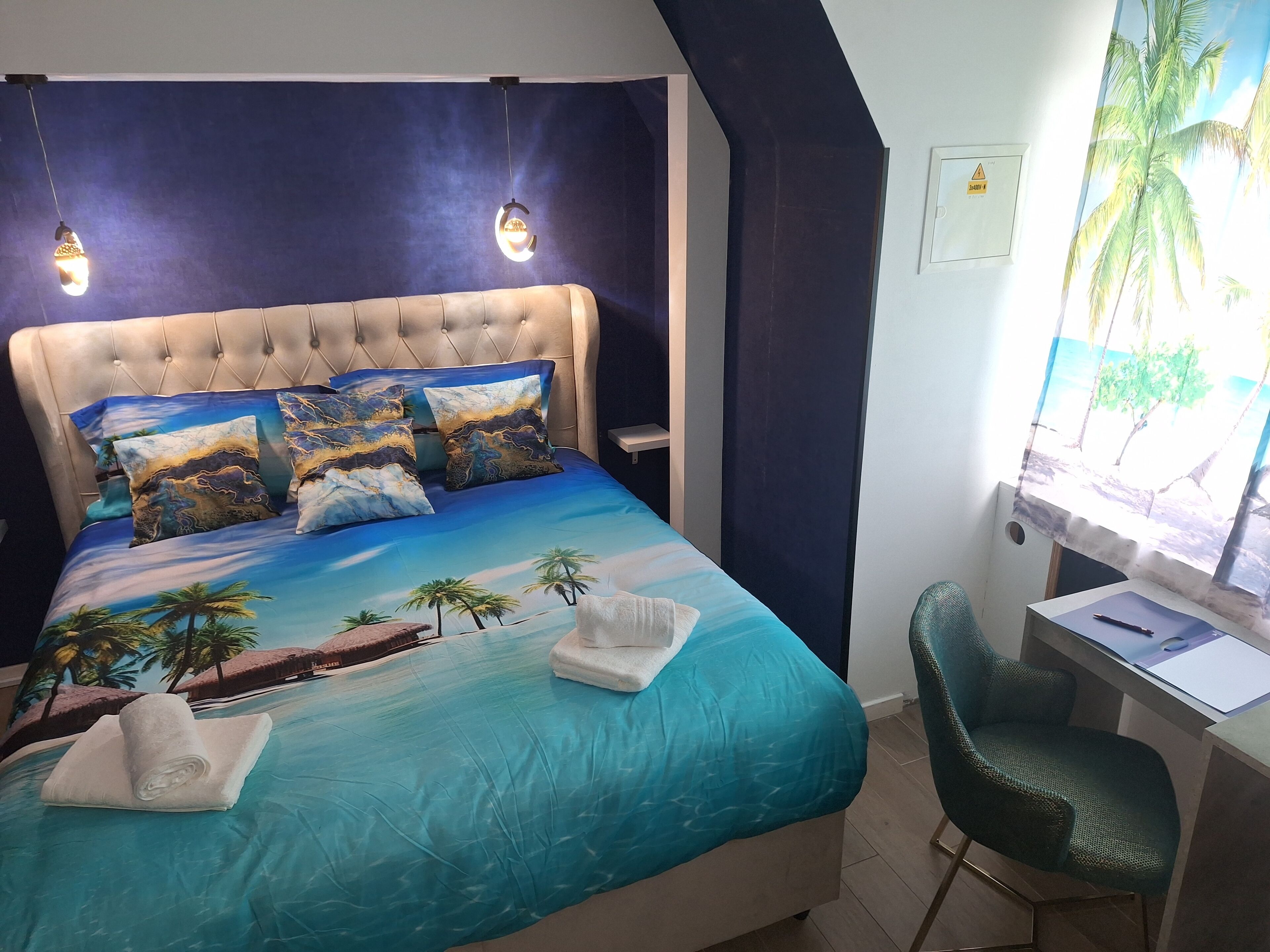 Un dormitorio con una cama decorada con un diseño de playa tropical, un escritorio con una silla y una ventana que ofrece vistas a palmeras.