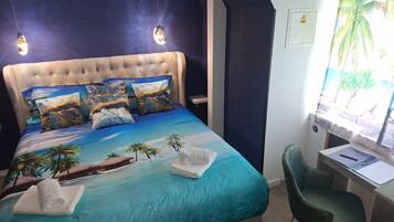 Un dormitorio con una cama decorada con un diseño de playa tropical, un escritorio con una silla y una ventana que ofrece vistas a palmeras.