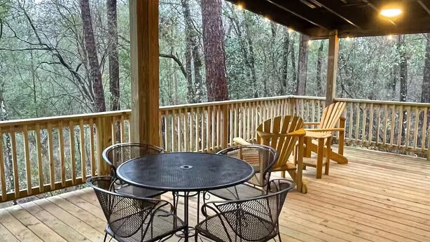 A covered outdoor deck with a round metal table and chairs, surrounded by a wooden railing and string lights. - Quiet, Creekside Home with Rain Spa Shower, Unplug & Relax (Collins)
