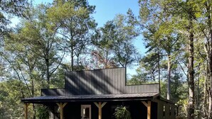A rustic cabin with a porch, surrounded by trees. - Quiet, Creekside Home with Rain Spa Shower, Unplug & Relax (Collins)