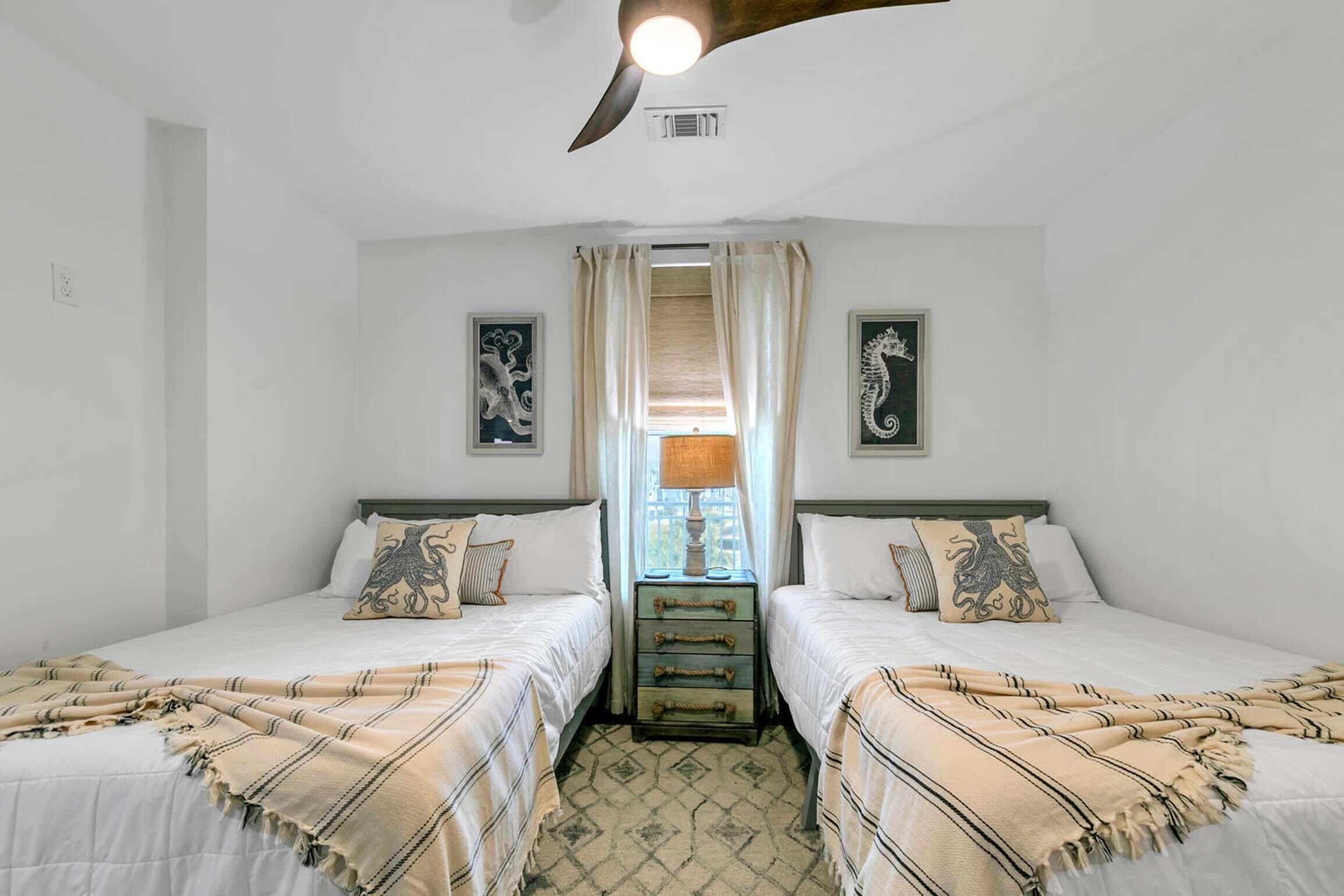 A room with two beds, a wooden chest of drawers, a ceiling fan, and two wall-mounted artworks.