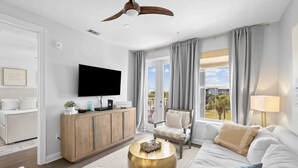 Ein modernes Wohnzimmer mit einem Flachbildfernseher, einer Holzkommode, einem runden Couchtisch aus Gold, einem weißen Sofa, einem beigen Sessel und Blick ins Freie durch große Fenster.