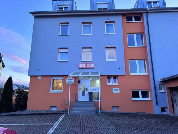 A multi-story hotel with a red and blue facade, a visible entrance with steps, and a sign indicating the name 'HOTEL SOAD'. - Hotel Soad (Salach)