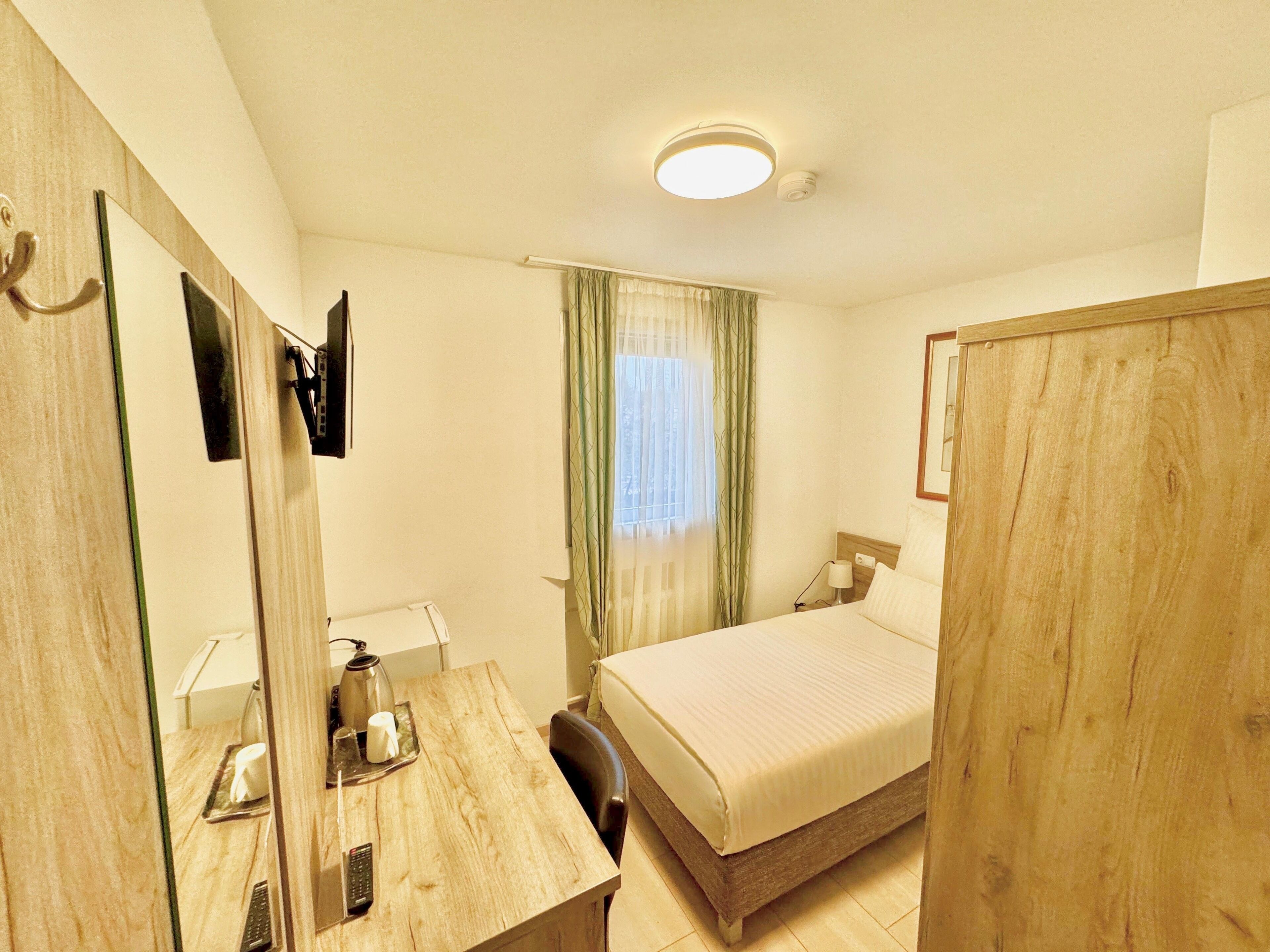 A hotel room with a bed, a desk with a lamp, a mirror, and a window with curtains.