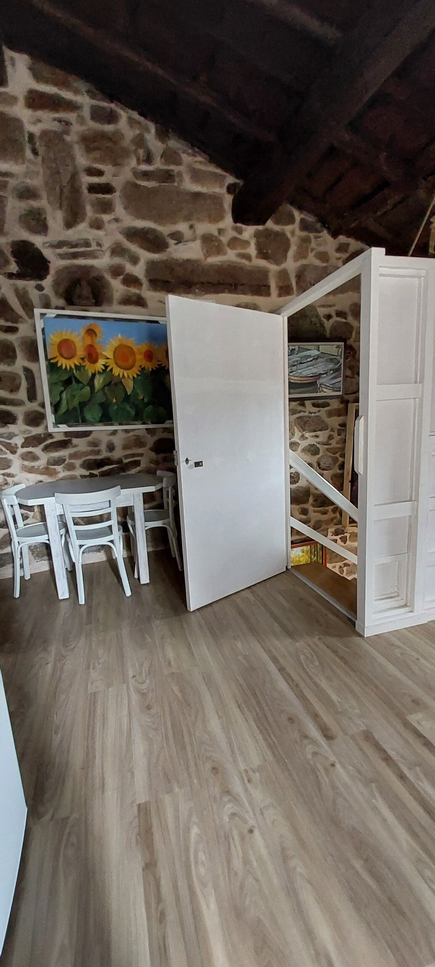 A room with a stone wall, a painting of sunflowers, a white door, a white wardrobe, and a small table with chairs.