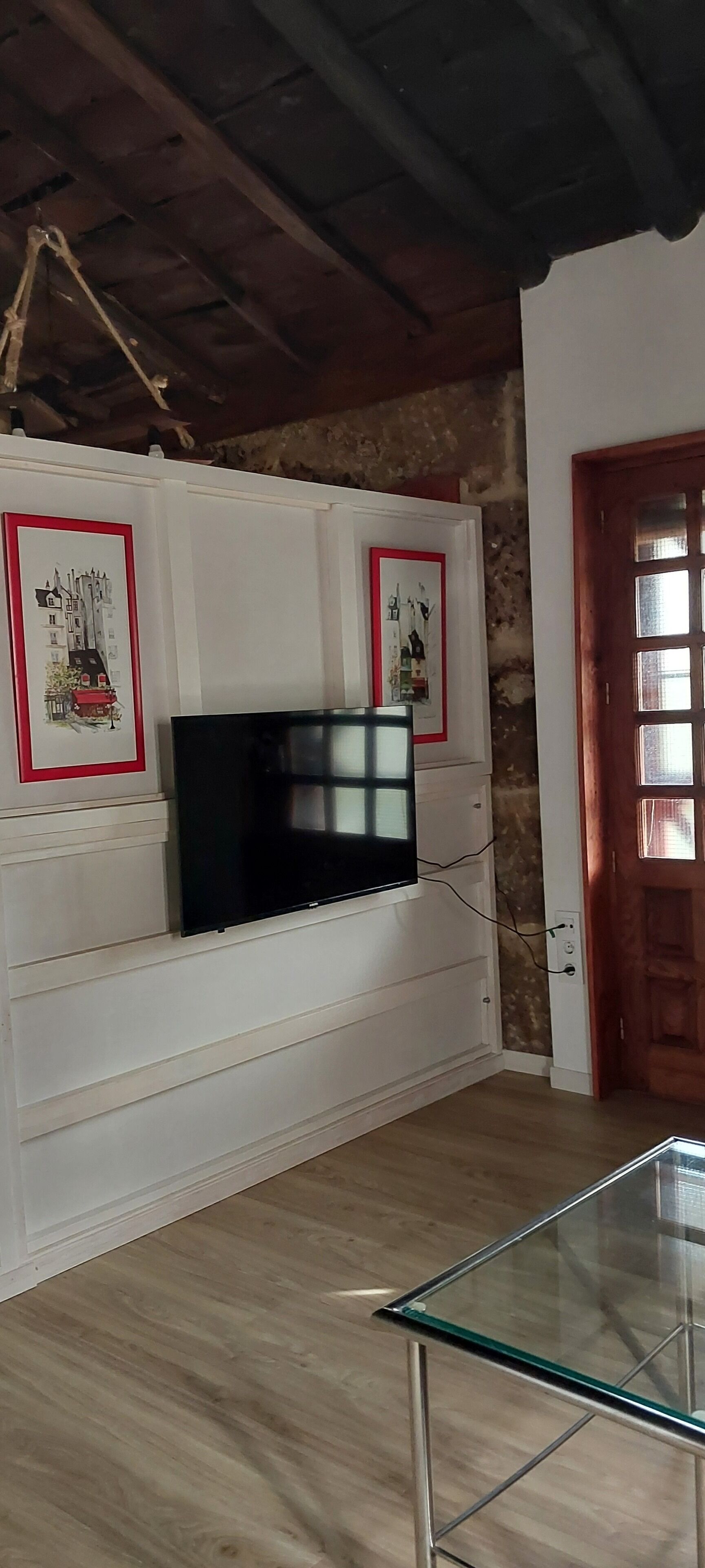 A room with a white built-in entertainment unit, a flat-screen TV, and framed pictures. There is a wooden door with glass panels and a glass-top table in the foreground.