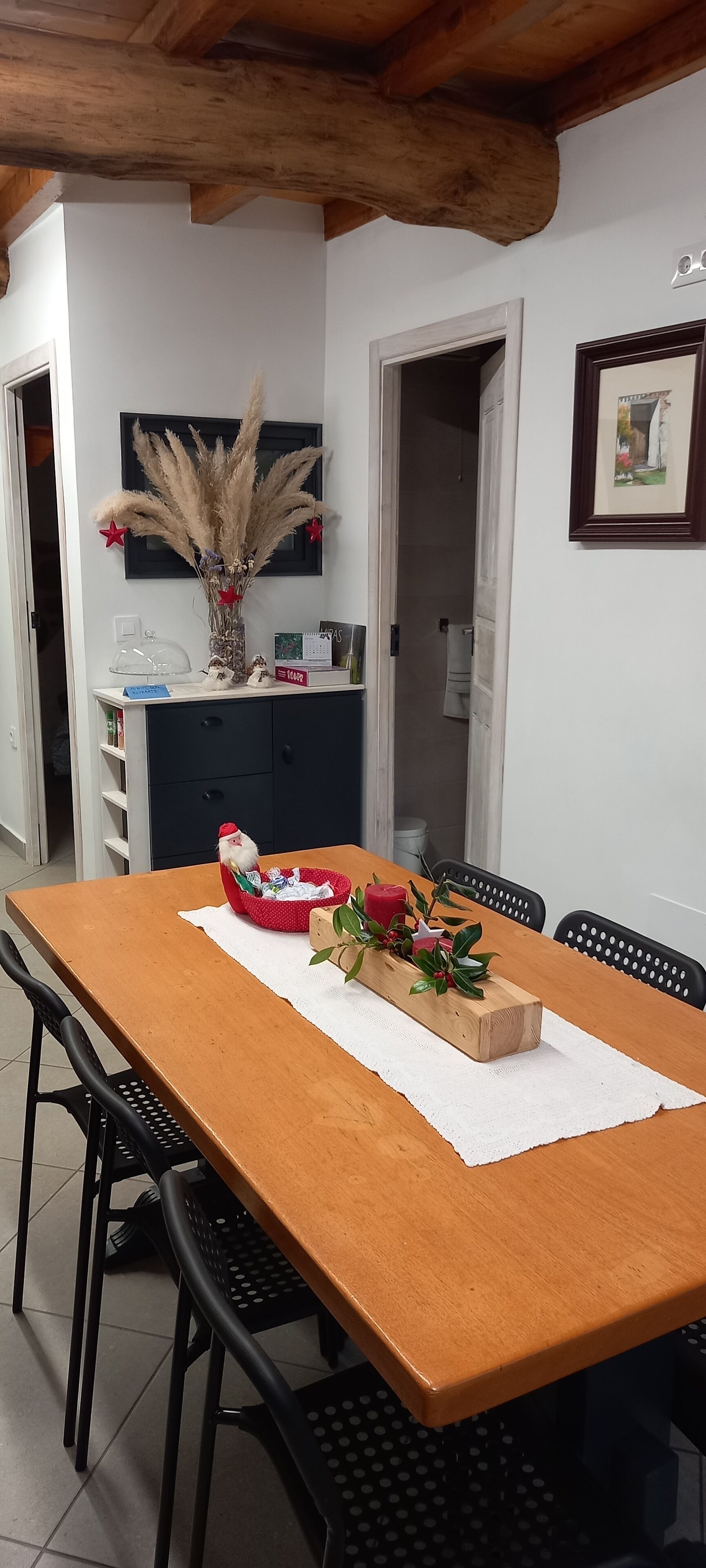 Un comedor con una mesa de madera, sillas negras, un florero con plantas secas y una cesta roja con gorro de Papá Noel.