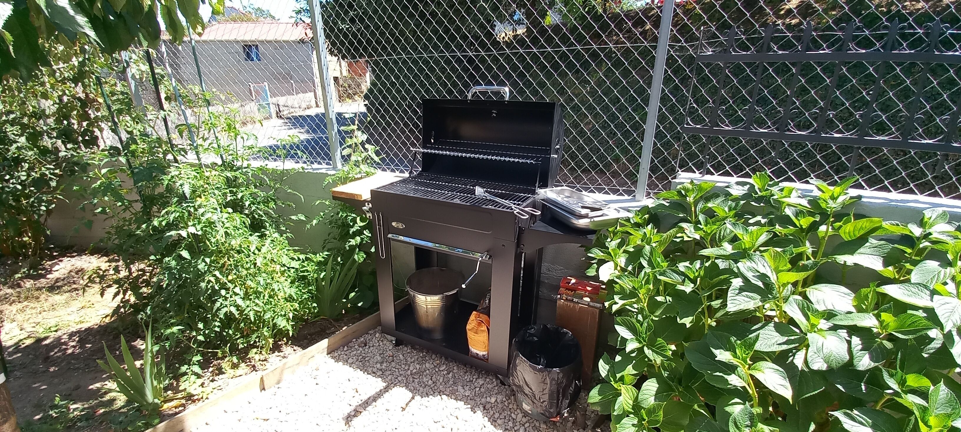 Un espace barbecue extérieur, délimité par une clôture en treillis métallique, comprenant un barbecue, une poubelle et des plantes en pot.