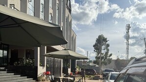 An outdoor seating area with tables and chairs, a covered patio, and a parking area adjacent to a building with multiple windows.