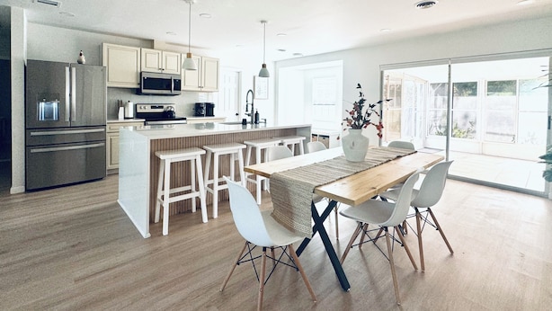 Una cocina moderna con una isla central, taburetes blancos tipo bar y una mesa de comedor con sillas.