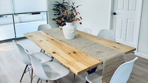 Un comedor con mesa de madera, sillas blancas, un florero con flores y un ventanal grande.