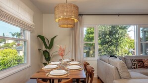 A dining area with a wooden table set for six, surrounded by chairs, a woven pendant light, and a view of greenery outside.