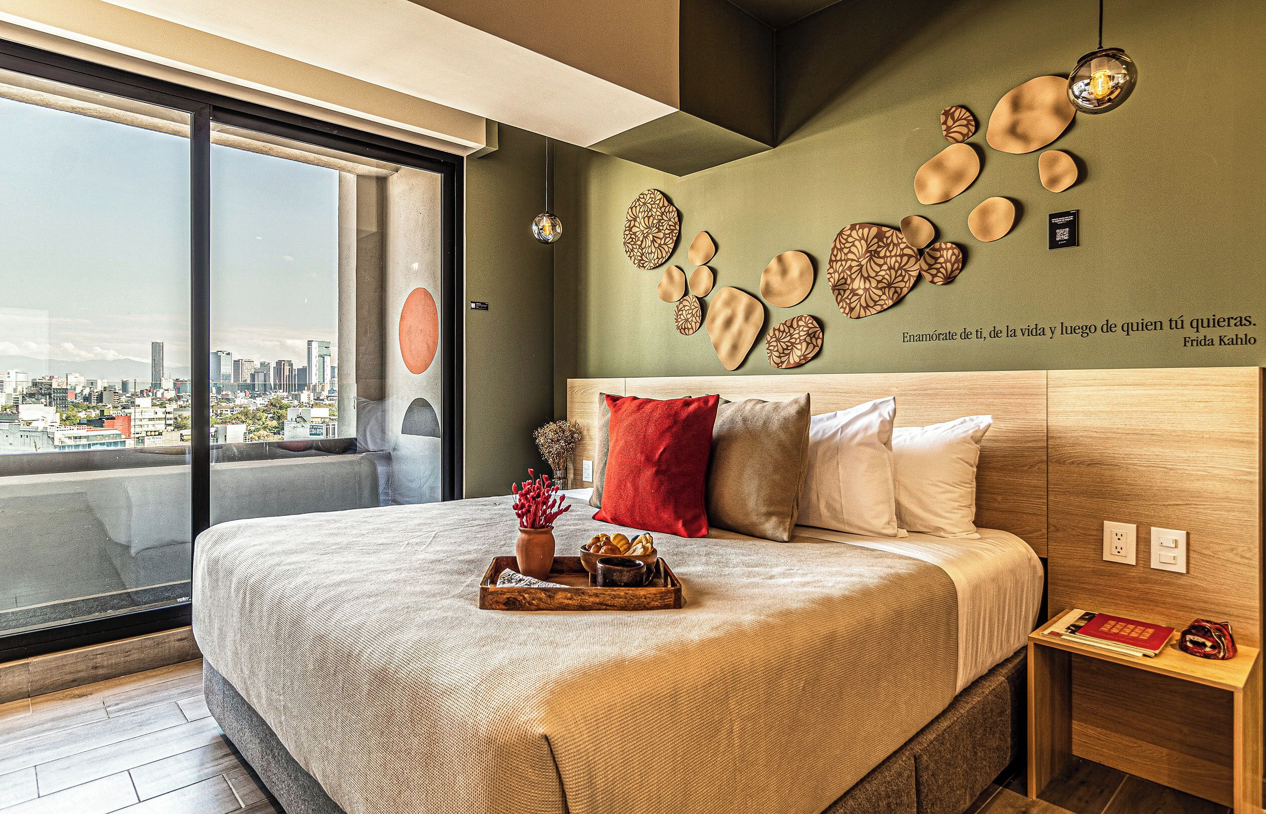 Un dormitorio moderno con vistas a la ciudad, una cama grande con muchas almohadas, decoración de pared y una mesita de noche con libros.