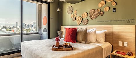 A modern bedroom with a city view, a large bed with multiple pillows, decorative wall art, and a bedside table with books.