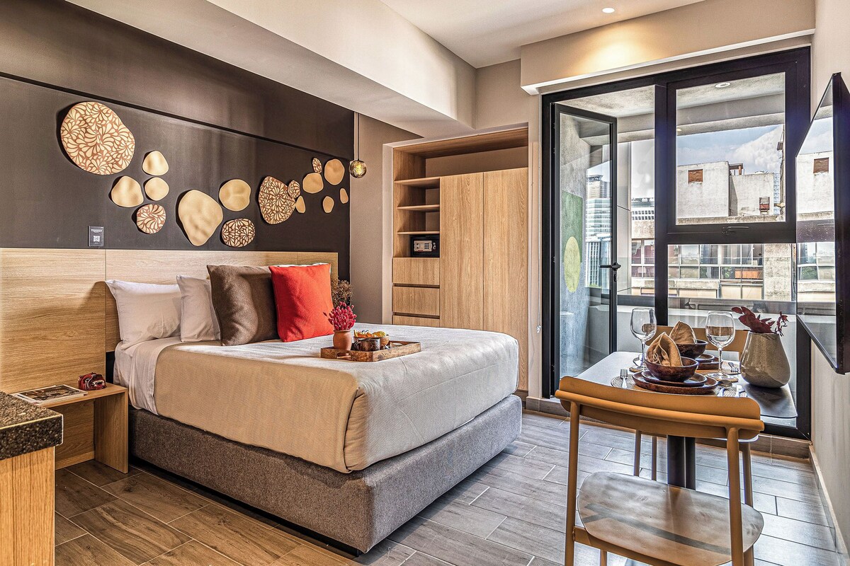 A modern hotel room with a large bed, a wooden headboard, a dining area with a table set for two, and a city view through a large window.