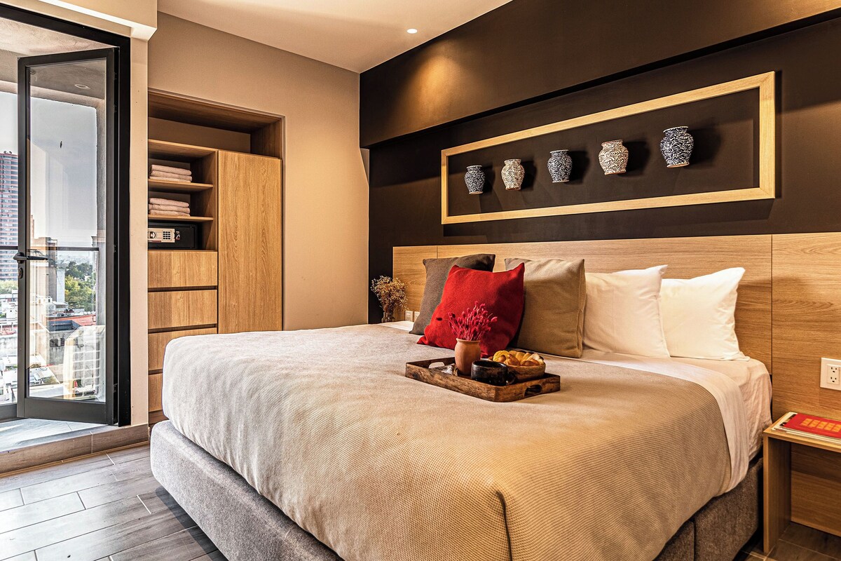 A modern bedroom with a large bed, a wooden wardrobe, a nightstand, and a city view through a glass door.