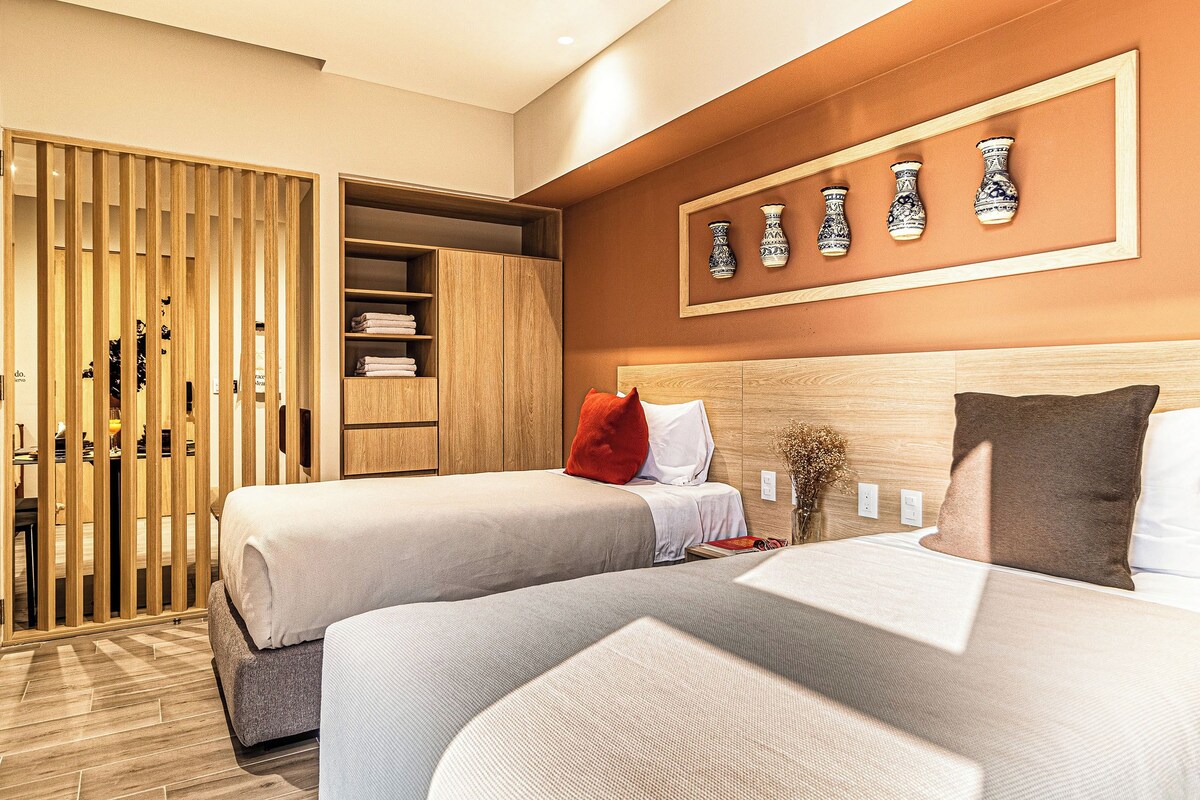 Une chambre d’hôtel avec deux lits, une armoire en bois, une étagère murale où sont exposés des vases, et une petite table sur laquelle se trouvent un vase et un livre.