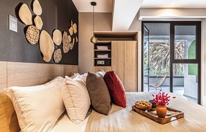 Une chambre à coucher moderne avec un lit, des oreillers, une table de chevet avec une lampe, une étagère murale avec des livres et une vue sur l’extérieur par une porte vitrée.